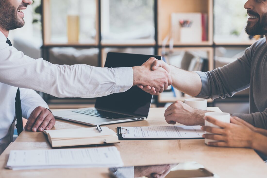 two men shaking hands over a table with a laptop