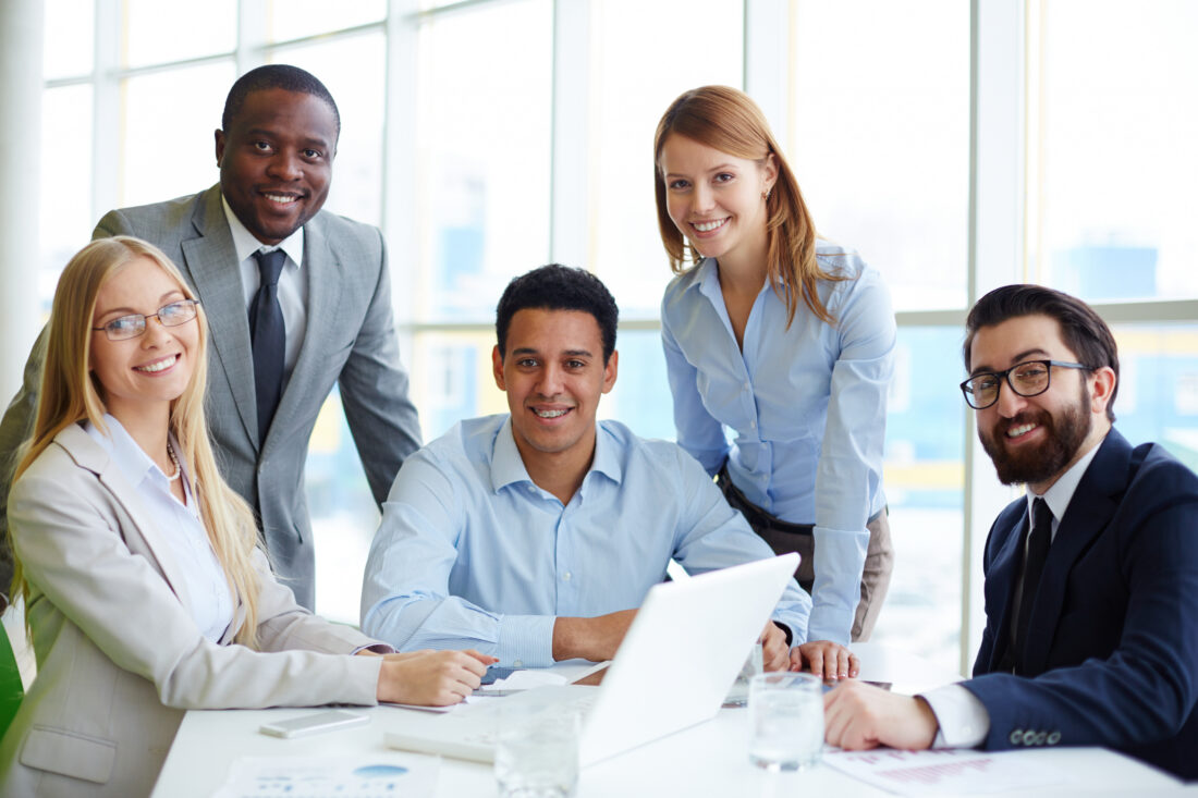 a group of business people sitting around a table with a laptop and smiling at the camera